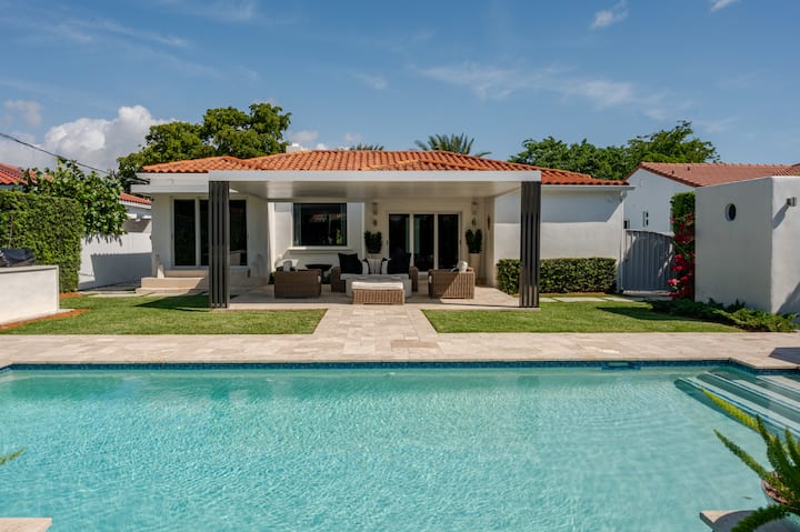Un espace extérieur sophistiqué avec une piscine, une terrasse spacieuse, des sièges en osier et une maison blanche avec des tuiles rouges, dans un cadre paysager luxuriant.