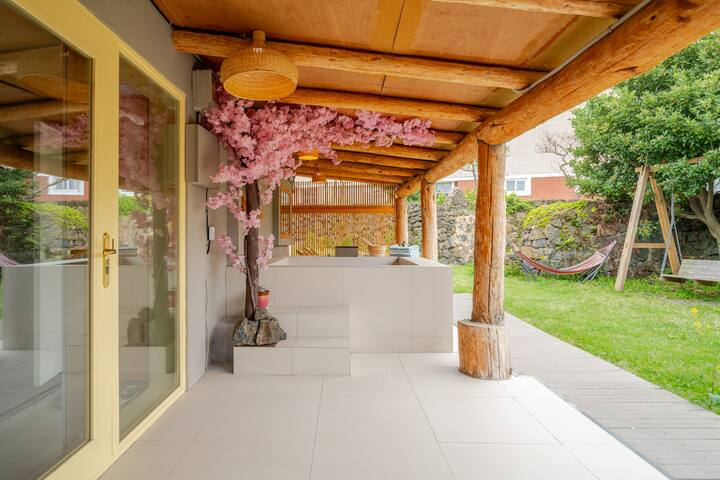 Terrasse couverte avec poutres en bois, arbre à fleurs roses, sol carrelé, ainsi qu'une balançoire et un hamac sur une pelouse à l'arrière-plan.