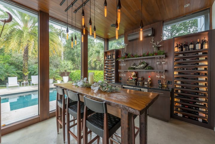 Un élégant bar intérieur avec des luminaires suspendus, des casiers à bouteilles et une vue sur la piscine encadrée par de grandes baies vitrées et des palmiers à l'extérieur.
