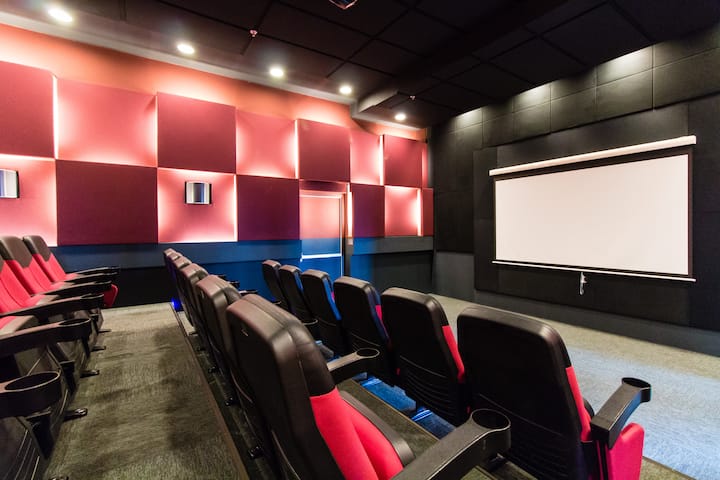 Un cinéma privé moderne avec des fauteuils inclinables rouge et noir, un écran de projection blanc et un éclairage mural chaleureux.