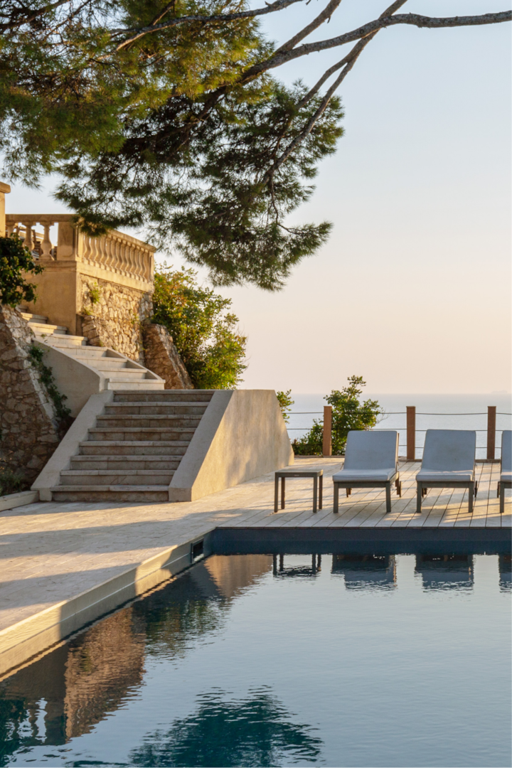Une piscine extérieure luxueuse avec des chaises longues, des marches en pierre menant à une terrasse et une vue sur l'océan, le tout encadré par des arbres au coucher du soleil.