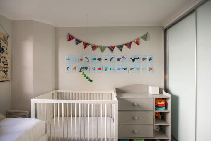 Une chambre de bébé confortable et bien éclairée avec un lit pour bébé, des décorations murales colorées, notamment des illustrations d'animaux et des fanions, ainsi qu'une commode grise remplie de jouets et d'articles pour bébé.