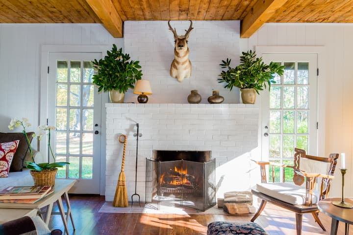 Salon avec un feu dans une cheminée en briques blanches, une tête de cerf au mur, des plantes et des meubles en bois. Les portes-fenêtres de chaque côté laissent entrer une lumière abondante, créant une atmosphère chaleureuse.