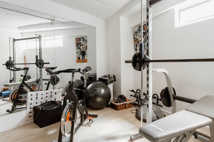Une salle de sport lumineuse avec des miroirs, un vélo d'appartement, un banc de musculation, une barre d'haltères, un ballon de yoga et des équipements de fitness rangés dans des paniers.