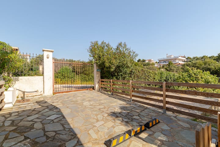 Une allée privée pavée en pierre fermée par un portail en métal et une clôture en bois, donnant sur la végétation et les maisons environnantes par une journée ensoleillée.