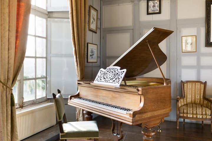 Une pièce baignée de lumière naturelle avec des œuvres d'art encadrées, une chaise vintage et un piano à queue en bois surmonté d'un pupitre à partitions orné près de grandes fenêtres habillées de rideaux.