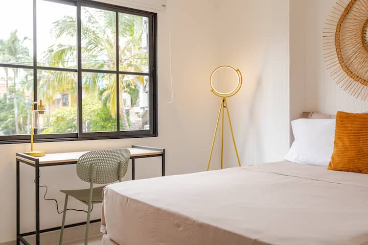 Une chambre confortable baignée de lumière naturelle, avec un bureau et une chaise près d'une fenêtre, un lit soigneusement fait et une élégante lampe dorée.