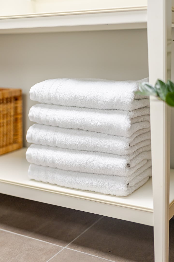 Quatre serviettes blanches pliées et disposées sur une étagère, à côté d'un panier en osier et d'une plante verte, soulignant la propreté et le confort des voyageurs.