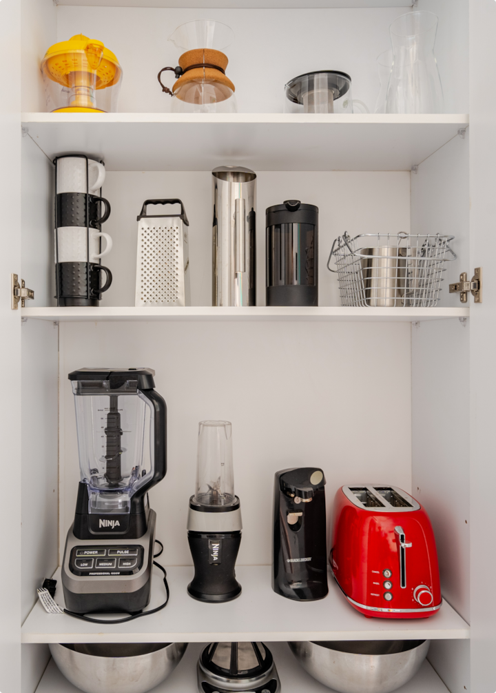 Une armoire bien organisée avec des appareils et des ustensiles de cuisine tels qu'un mixeur, une cafetière à piston, un moulin à café, un grille-pain, des tasses et des récipients en verre.