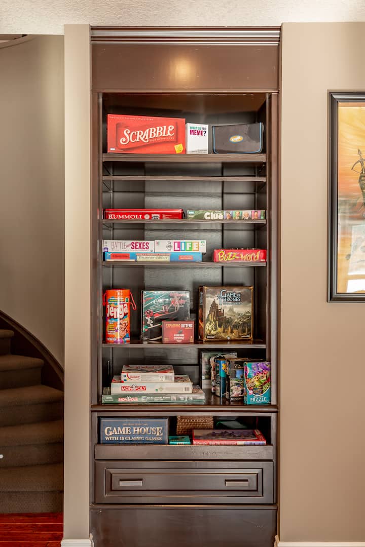 Une grande bibliothèque remplie de jeux de société tels que Jenga, Monopoly ou Scrabble, ainsi que des puzzles, mettant en avant des activités à faire en famille dans un espace de vie chaleureux.