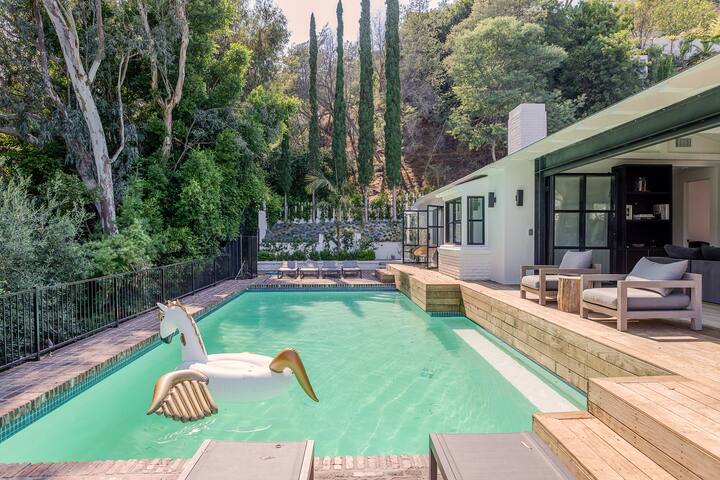 Une piscine turquoise avec une bouée licorne, bordée d'une terrasse en bois et de chaises longues. La piscine est entourée d'une végétation dense, de grands cyprès et d'une maison blanche moderne avec de grandes fenêtres.
