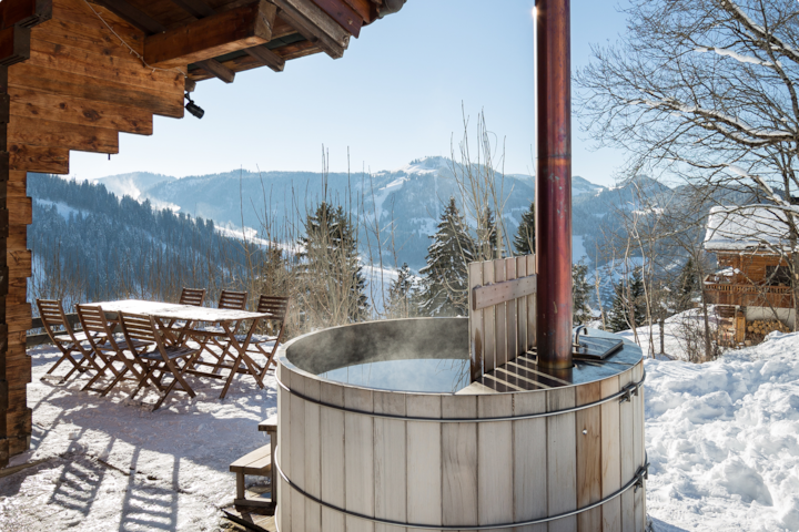 Un cadre extérieur enneigé montrant un jacuzzi en bois rustique, une table à manger en bois avec des chaises et une vue pittoresque sur les montagnes et les pins.