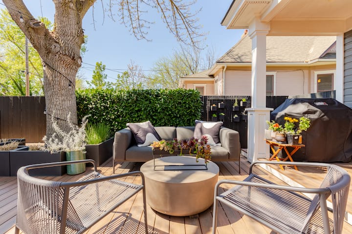 Une terrasse en bois ensoleillée avec des sièges, des plantes en pot et un barbecue, ombragée par un grand arbre et bordée de feuillages.