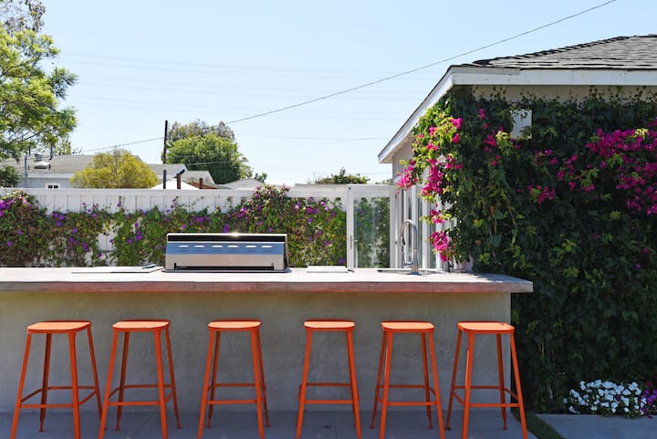 Un bar extérieur équipé d'un barbecue, d'un évier et de tabourets orange, avec un mur fleuri et un ciel dégagé en arrière-plan.