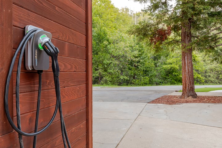 Borne de recharge pour véhicules électriques montée sur un mur en bois, entourée d'une végétation luxuriante et d'un arbre en arrière-plan.