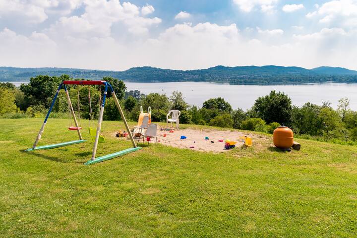 Une aire de jeux dans un jardin avec une balançoire, un bac à sable et des jouets sur le sol, surplombant un lac paisible et des collines boisées.
