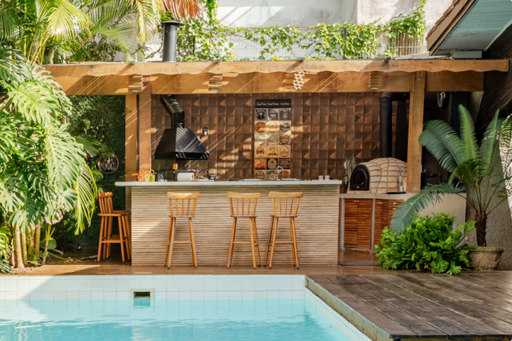 Un espace piscine paisible avec une végétation luxuriante, un bar au bord de la piscine avec des sièges et une terrasse en bois qui s'étend le long de l'eau.