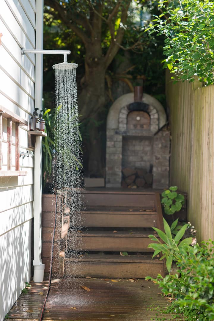 Un jardin ensoleillé et entouré de feuillage, avec une douche extérieure fixée à un mur blanc et des marches menant à un four à pizza en pierre.