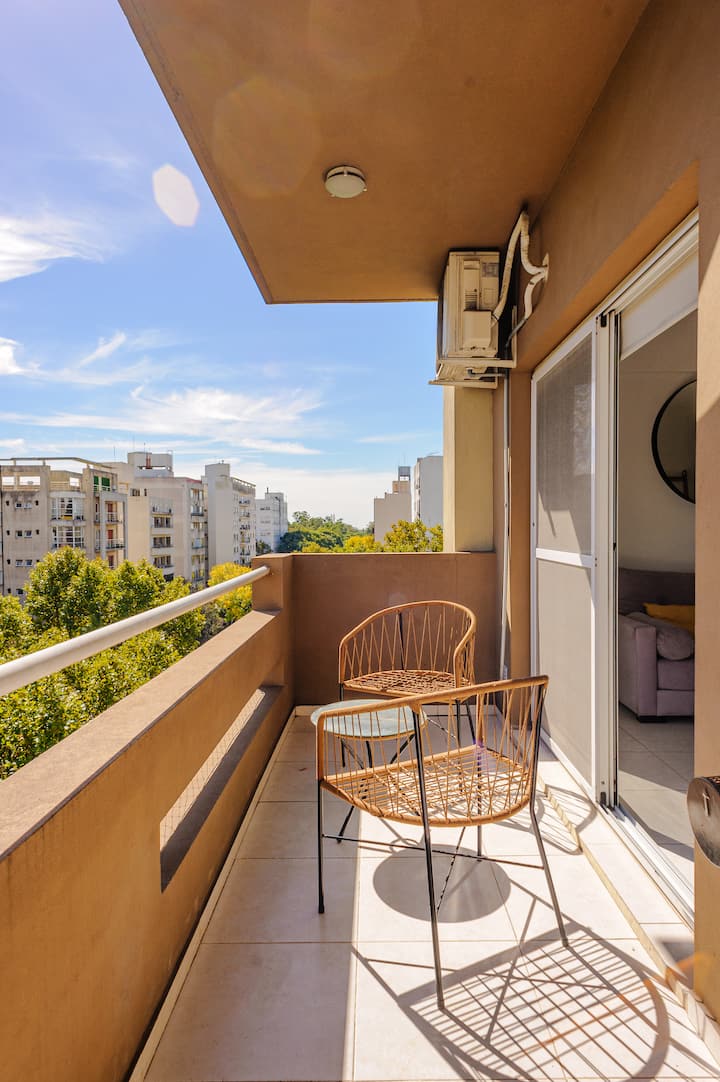 Balcon avec murs beiges, deux chaises minimalistes et vue sur des arbres et des bâtiments résidentiels par une journée ensoleillée.