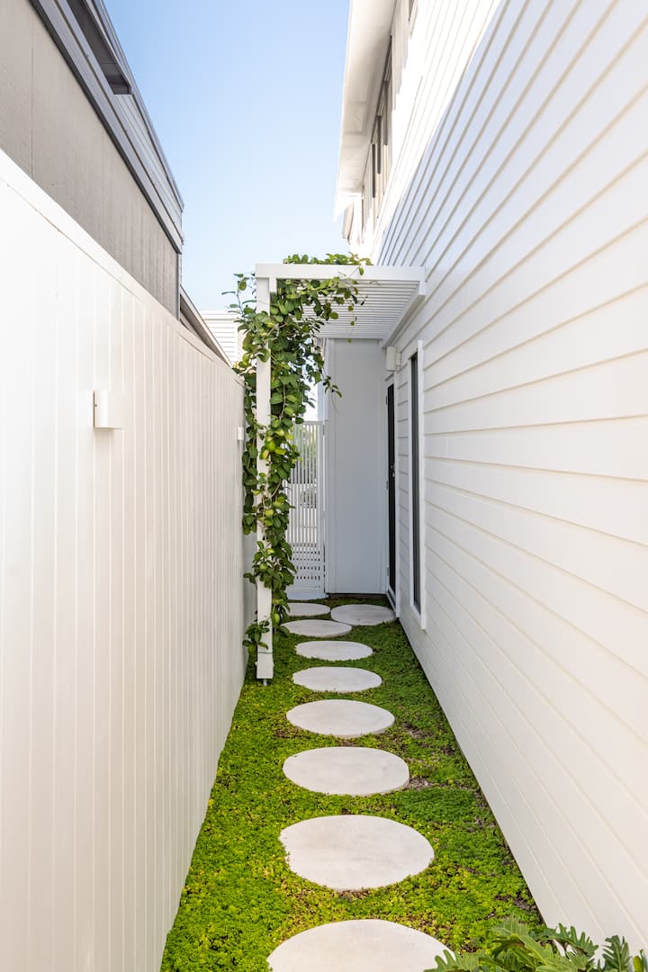 Un chemin extérieur avec des pierres plates posées sur la verdure, encadré par des murs blancs, et une pergola avec des vignes menant à un portail.