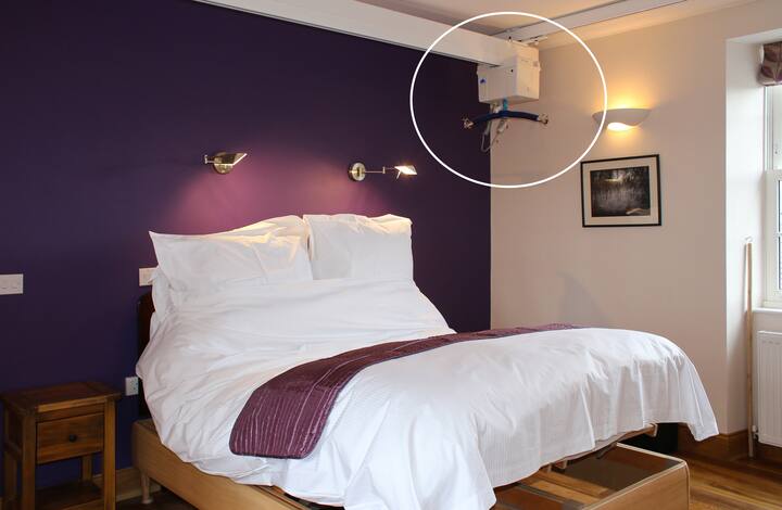 Une chambre accessible avec un mur violet, des draps blancs et un lève-personne plafonnier. Une table de chevet en bois et des tableaux sont également visibles.