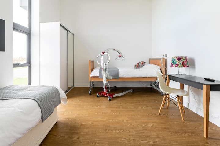 Une chambre lumineuse et accessible avec un lit médicalisé et un lève-personne à côté, un bureau avec une chaise et de grandes fenêtres offrant une lumière naturelle.