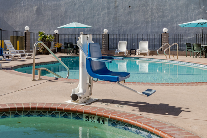 Un espace piscine avec un jacuzzi au premier plan doté d'un lève-personne avec un siège bleu, et des chaises longues et parasols en arrière-plan.