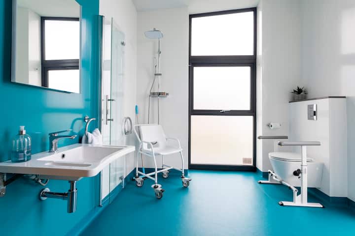 Une salle de bain accessible avec une douche de plain-pied équipée d'une chaise, de barres d'appui autour des toilettes et d'un lavabo abaissé pour un accès facile.