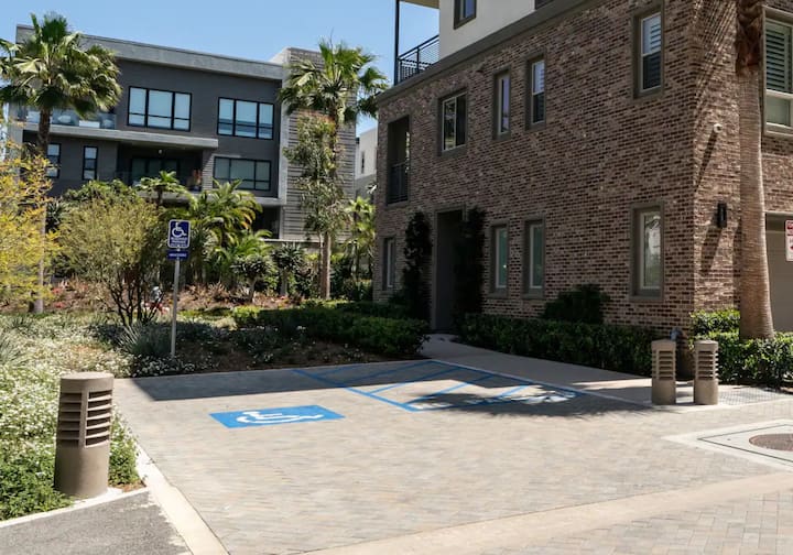 Deux places de stationnement accessibles avec des symboles peints sont situées à côté d'un bâtiment en briques, entouré de plantes et d'arbres.