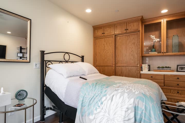 Une chambre accessible et bien éclairée avec un lit réglable et des draps à fleurs, une table d'appoint avec des produits de base et des meubles en bois avec des objets de décoration en arrière-plan.