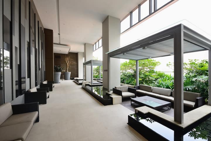 Un espace lounge d'immeuble ouvert et lumineux avec des sièges sous des auvents métalliques, des aménagements paysagers et des jeux d'eau décoratifs.