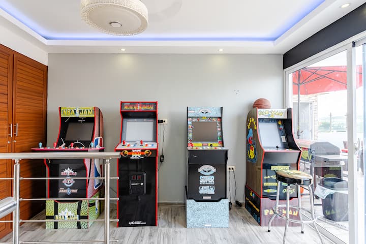 Un espace de jeu soigné équipé de 4 bornes d'arcade vintage proposant des jeux populaires comme Mortal Kombat et Pac-Man, à côté d'une porte vitrée qui révèle une terrasse avec salon d'extérieur.
