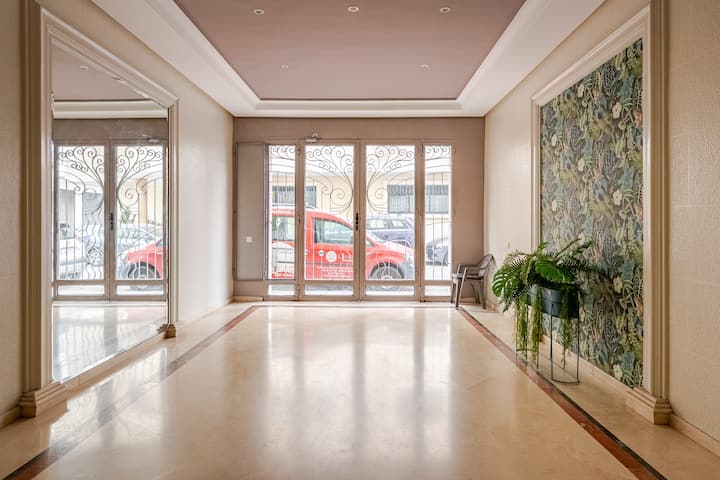 Hall d'entrée spacieux d'un immeuble d'appartements avec des sols cirés, de grands miroirs décoratifs, des fenêtres en fer forgé et un mur décoré de papier peint floral tropical.