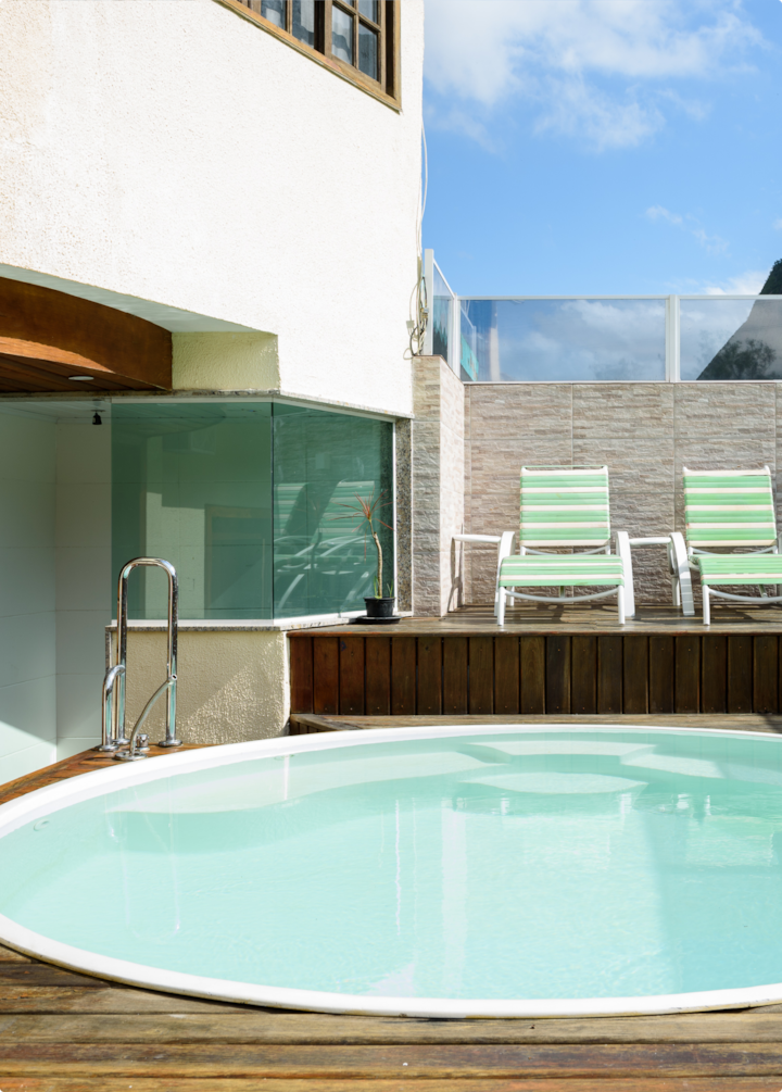 Un patio ensoleillé avec une petite piscine ronde, des chaises longues vert et blanc, et une barrière en verre donnant sur des montagnes pittoresques.