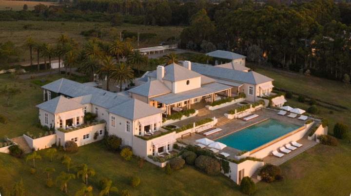 Perspective aérienne d'un vaste domaine au crépuscule. Elle met en valeur une pelouse bien entretenue, des palmiers, une piscine et de nombreux équipements extérieurs.
