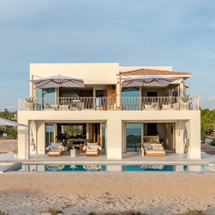 Une maison de deux étages au design minimaliste. Elle est dotée de balcons avec des sièges à l'ombre, d'une piscine et de vues panoramiques sur la végétation et le ciel.