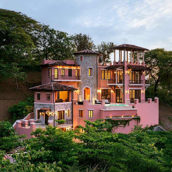 Une villa lumineuse et entourée d'une végétation luxuriante, avec une façade rose et en pierre. Elle est dotée de balcons, de grandes fenêtres et d'un espace piscine éclairé au crépuscule.