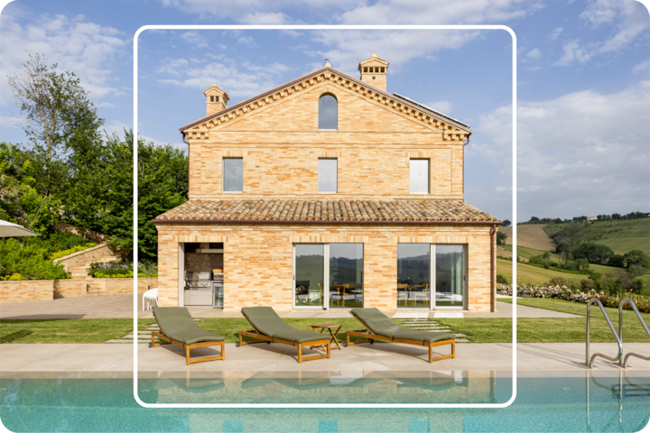 Une grande villa en briques sur fond de collines verdoyantes et d'un ciel dégagé. Des chaises longues se trouvent au bord d'une piscine.