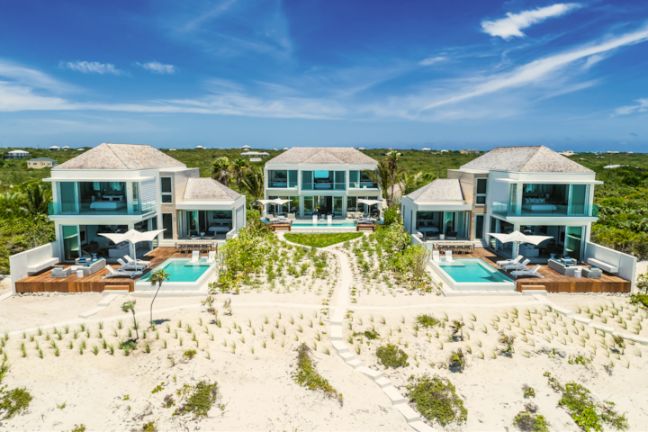 Photo réalisée par un drone de villas modernes agrémentées de balcons en verre, de piscines privées et d'aménagements paysagers. L'arrière-plan est composé d'un terrain sablonneux et d'un ciel bleu vif.