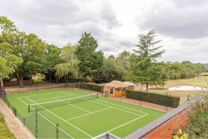 Cliché réalisé par un drone d'une propriété dotée d'un court de tennis privé. Il est entouré d'arbres, d'un mur en briques et d'une petite cabane à proximité, avec un étang pittoresque en arrière-plan.