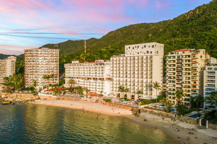 Plan large dévoilant un bâtiment en bord de mer, avec des collines en arrière-plan, un rivage sablonneux, des baigneurs et un ciel du soir coloré.