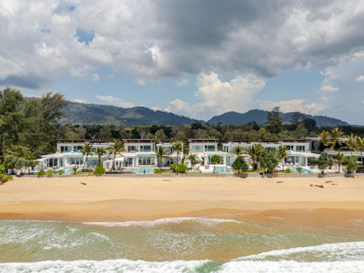 Vue d'en haut de logements côtiers élégants et dotés de piscines. En arrière-plan, il y a des palmiers, des montagnes et un ciel partiellement nuageux.