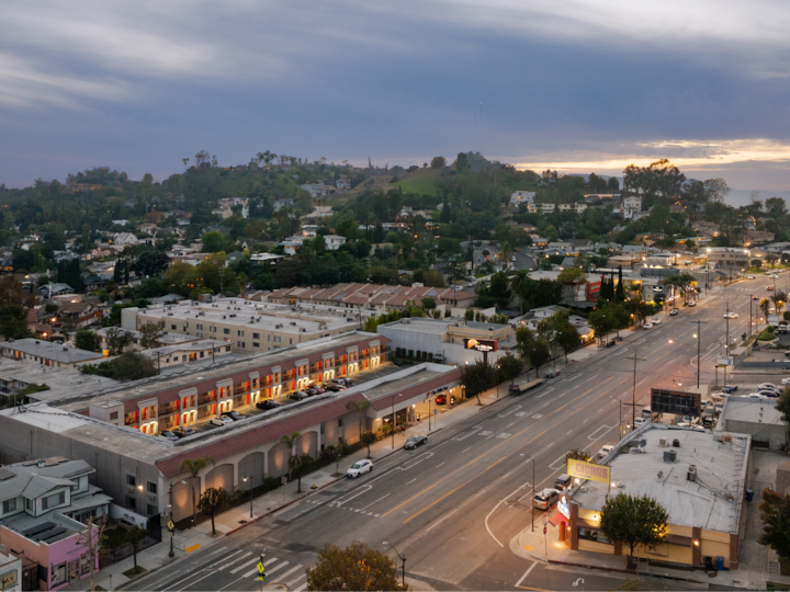 Vue aérienne d'un quartier au coucher du soleil. Sa rue principale est bordée de magasins éclairés, d'habitations et de végétation en arrière-plan.