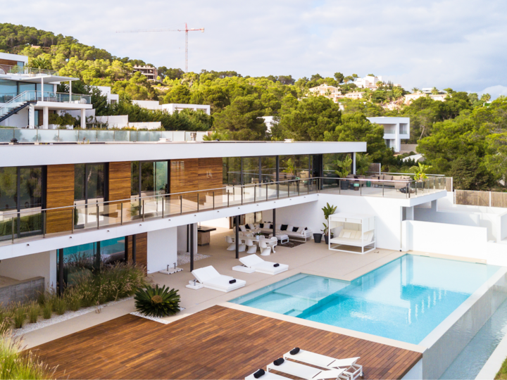 Une villa moderne sur deux étages. Elle est dotée d'une grande terrasse extérieure, d'une piscine à débordement et de chaises longues. Une luxuriante végétation l'entoure.