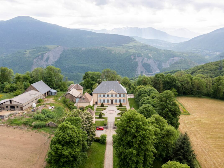 Vue aérienne d'un vaste domaine avec en son centre, une grande demeure. Des bâtiments, des jardins et des champs se trouvent à proximité, le tout sur fond de montagnes vallonnées.