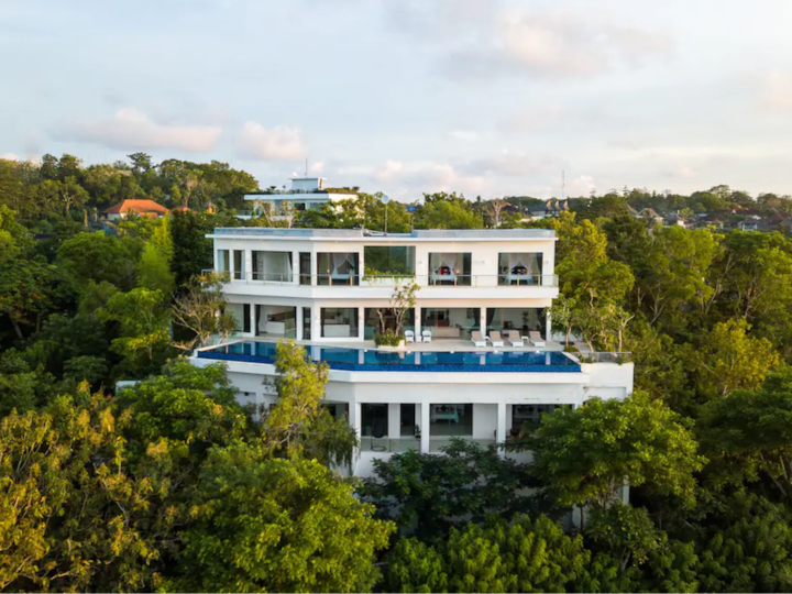 Vue aérienne d'une luxueuse villa blanche nichée au cœur d'une végétation dense. Le cliché met en valeur des terrasses ouvertes et une piscine bleue.