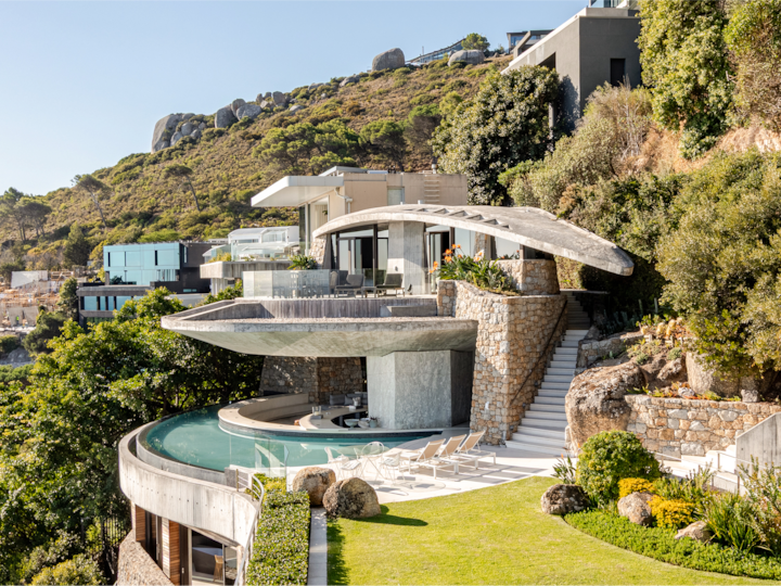 Une maison moderne et incurvée à flanc de coteau. Nichée sur un terrain rocheux, elle est dotée d'une piscine, de chaises longues et d'une végétation luxuriante.
