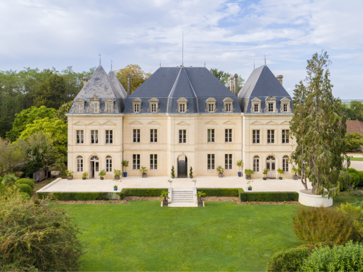 Vue aérienne d'un grand château élégant à l'architecture symétrique. Il se trouve au cœur d'un immense parc verdoyant, avec des arbres en arrière-plan.