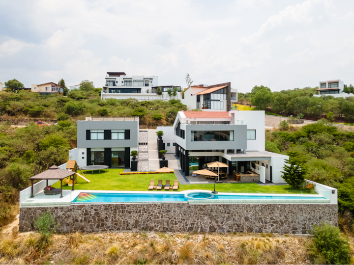 Maison d'architecte moderne et élégante, avec une grande piscine, un patio ombragé et une pelouse qui surplombe une végétation luxuriante depuis une colline surélevée.