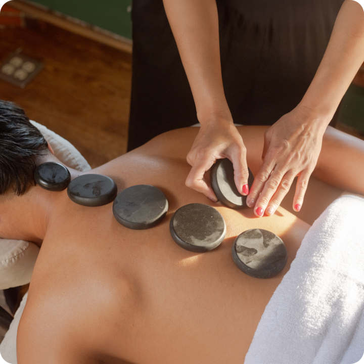Une personne reçoit un massage aux pierres chaudes relaxant ; une masseuse pose des pierres noires lisses sur son dos dans un cadre chaleureux et serein.